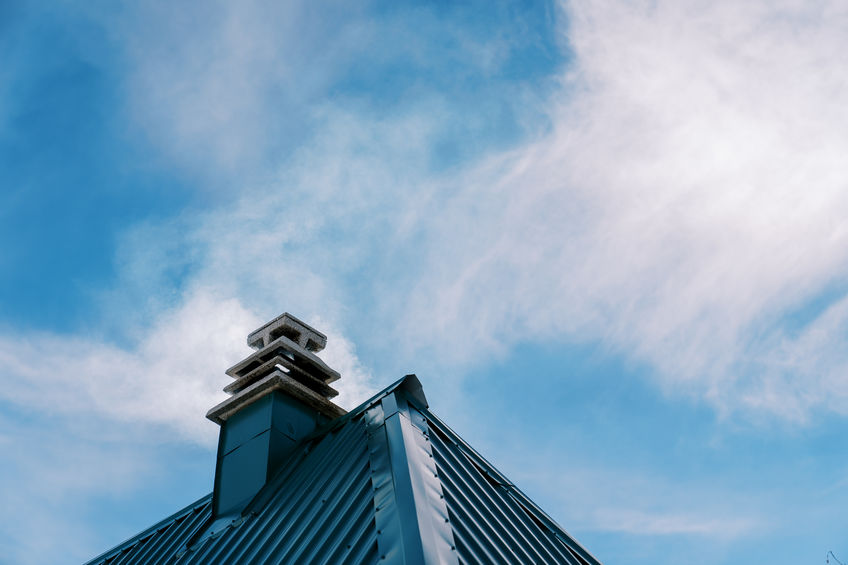 Smoke rises from a chimney on the green corrugated roof of a house against the blue sky. High quality photo